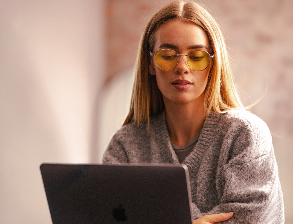 Woman wearing yellow-tinted blue light glasses working at a laptop in warm natural light